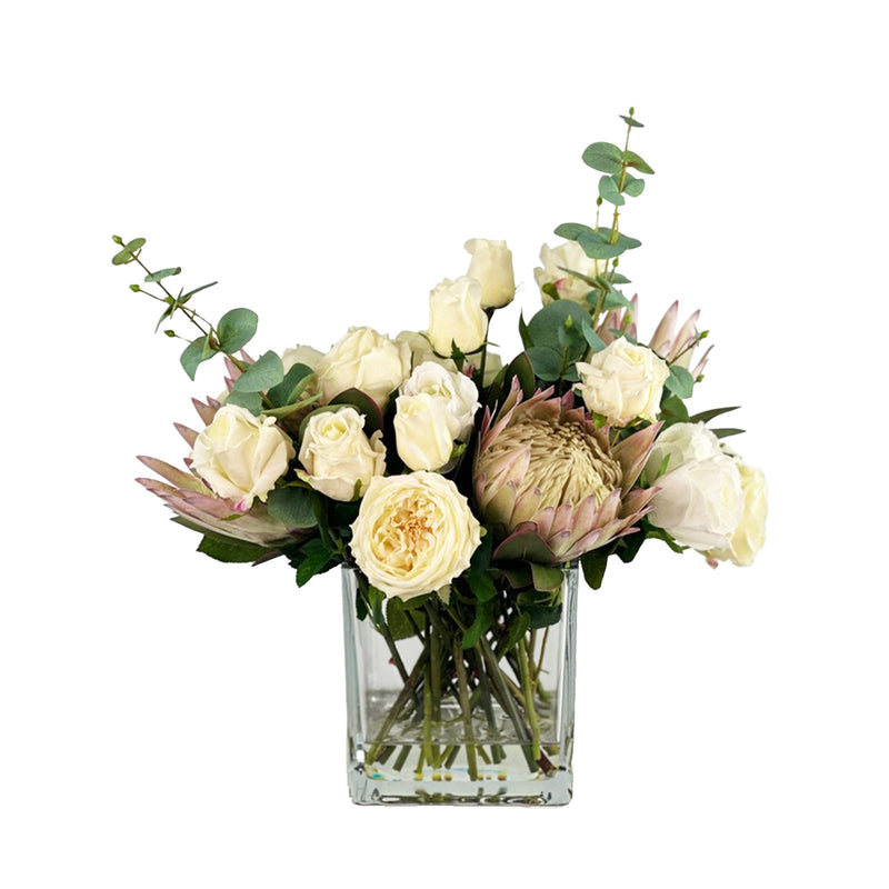 Bouquet of faux flowers with proteas and white roses in a clear vase with faux water on a white background